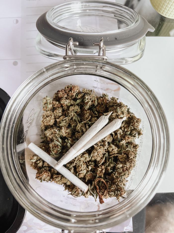 Glass jar filled with dried cannabis buds and rolled joints, viewed from above.